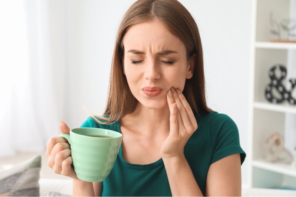 Woman holding a hot drink and wincing from tooth sensitivity or jaw pain